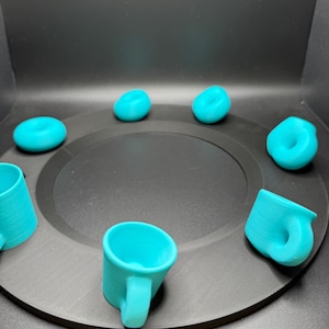 Op de afbeelding: Een verzameling turquoise 3D-geprinte mokken en donutvormige objecten, gerangschikt op een zwart cirkelvormig platform. De mokken hebben handvatten en afgeronde bodems. De donuts hebben een gladde, afgeronde vorm. De objecten zijn gelijkmatig verdeeld.