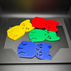 May include: Five colorful, bear-shaped objects in yellow, red, green, and blue are arranged on a dark surface. Each bear has cut-out eyes and a small mouth. The objects appear to be made of plastic or a similar material.