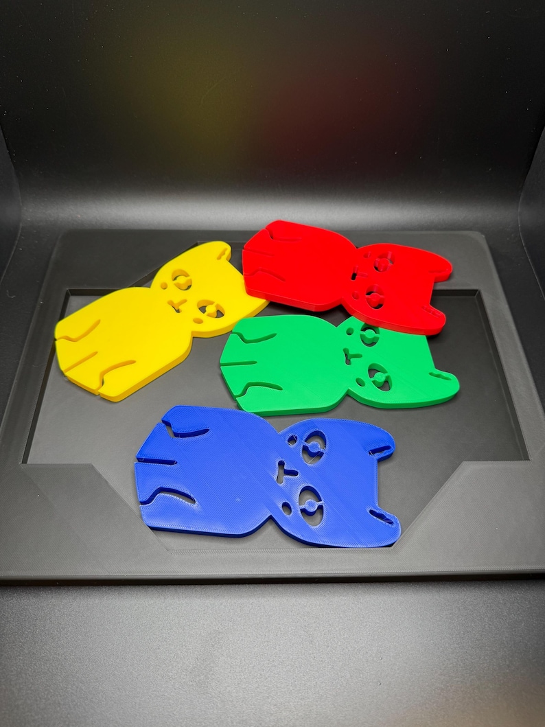 May include: Five colorful, bear-shaped objects in yellow, red, green, and blue are arranged on a dark surface. Each bear has cut-out eyes and a small mouth. The objects appear to be made of plastic or a similar material.