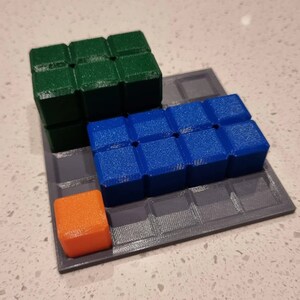 May include: A gray square game board with a green, blue, and orange cube on top. The cubes are arranged in a staggered pattern.