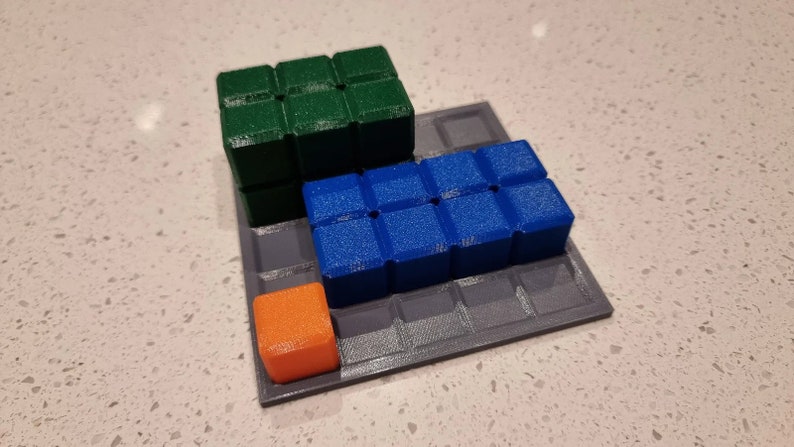 May include: A gray square game board with a green, blue, and orange cube on top. The cubes are arranged in a staggered pattern.