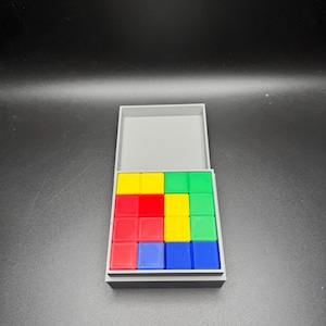 Puede incluir: Una caja gris con una tapa abierta, que revela una cuadr&iacute;cula de 4x4 bloques de colores. Los bloques son rojos, amarillos, verdes y azules.