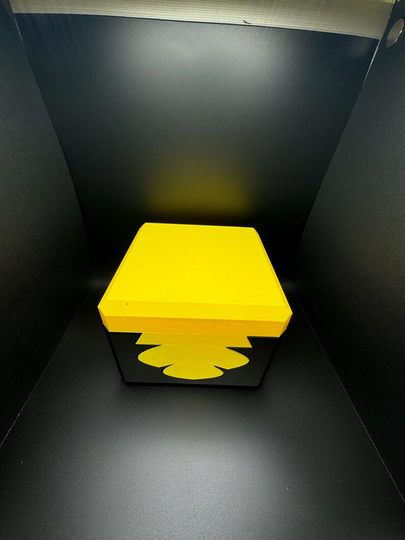 May include: A yellow square box with a black interior. The box has a simple design and is made of a sturdy material.