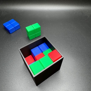 May include: A black square box filled with colorful, interlocking cube blocks. The blocks are red, green, and blue. Two additional blocks, one blue and one green, are positioned outside the box on a dark surface.