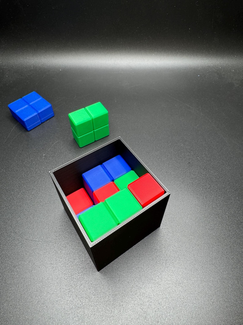 May include: A black square box filled with colorful, interlocking cube blocks. The blocks are red, green, and blue. Two additional blocks, one blue and one green, are positioned outside the box on a dark surface.