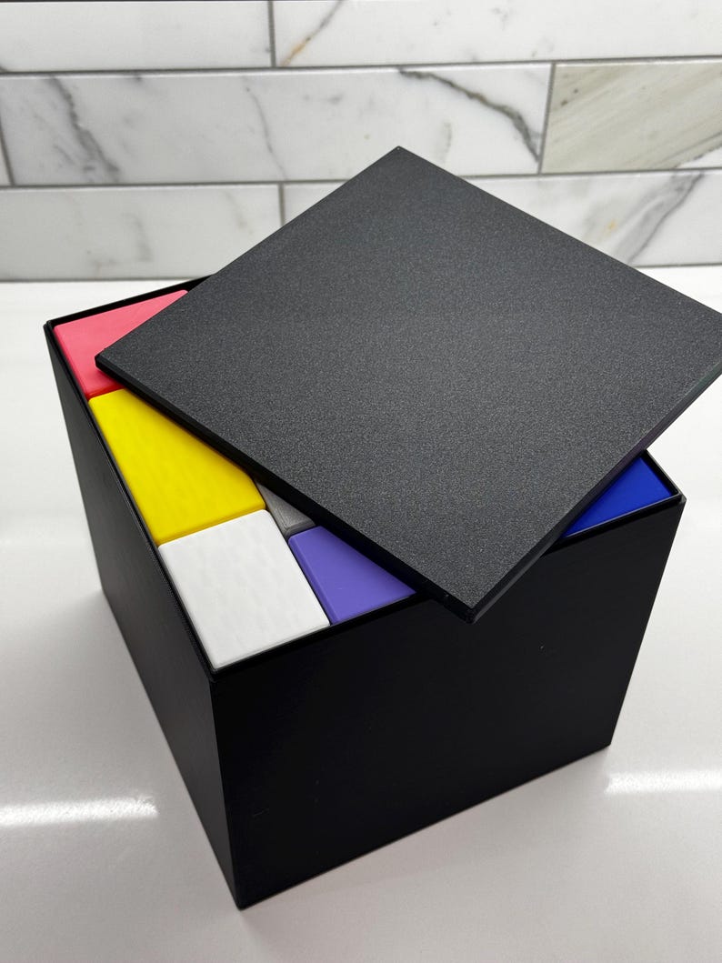 May include: A black box with a lid open, revealing a stack of colorful blocks. The blocks are red, yellow, white, purple, and blue.