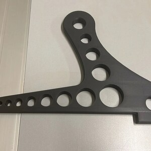 Może przedstawiać: Czarny metalowy wspornik wahacza motocyklowego z wieloma okrągłymi otworami.