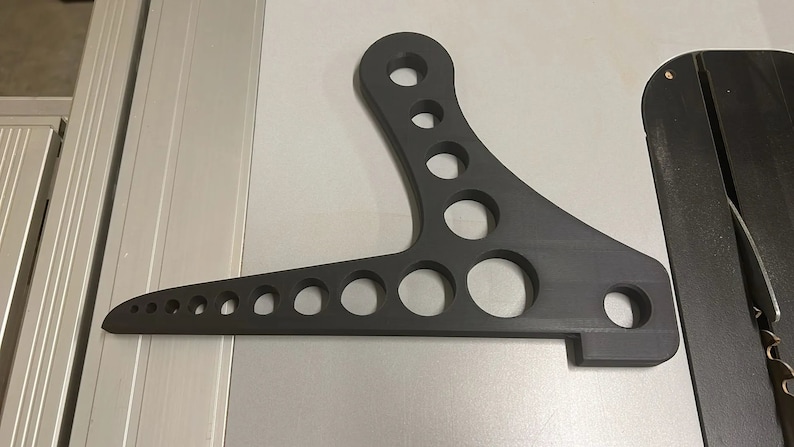 Może przedstawiać: Czarny metalowy wspornik wahacza motocyklowego z wieloma okrągłymi otworami.