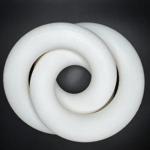 May include: A white, intertwined, circular sculpture against a black background. The sculpture appears to be made of a solid, smooth material, with a continuous, looping design. The form resembles a figure eight or a Mobius strip.