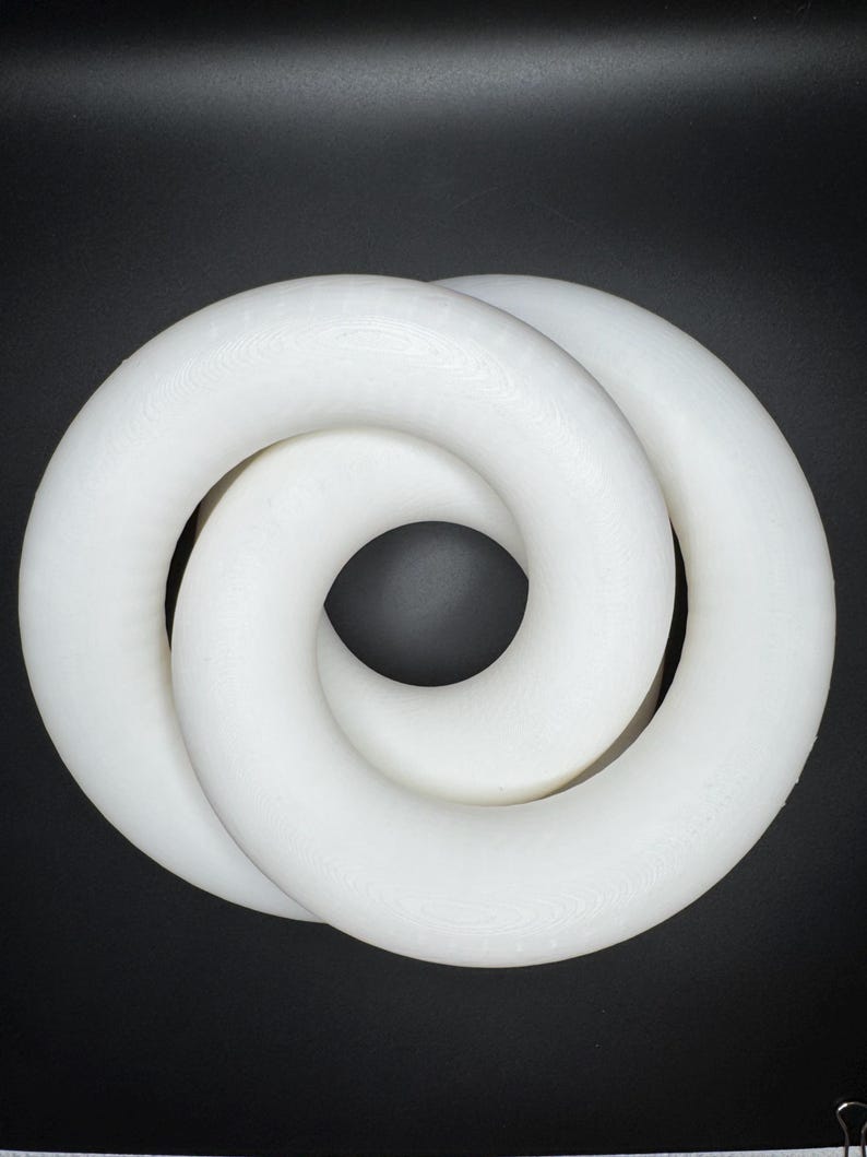 May include: A white, intertwined, circular sculpture against a black background. The sculpture appears to be made of a solid, smooth material, with a continuous, looping design. The form resembles a figure eight or a Mobius strip.