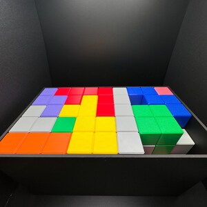 Op de afbeelding: Een kleurrijke puzzel gemaakt van kleine vierkante blokken in een houten doos. De blokken zijn in een patroon gerangschikt met verschillende kleuren, waaronder rood, blauw, groen, geel, oranje en grijs.