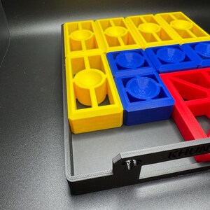 Puede incluir: Un tablero de juego negro con piezas de juego amarillas, azules y rojas. El tablero tiene un asa en el lateral y la palabra "KHUN" impresa en la parte delantera.