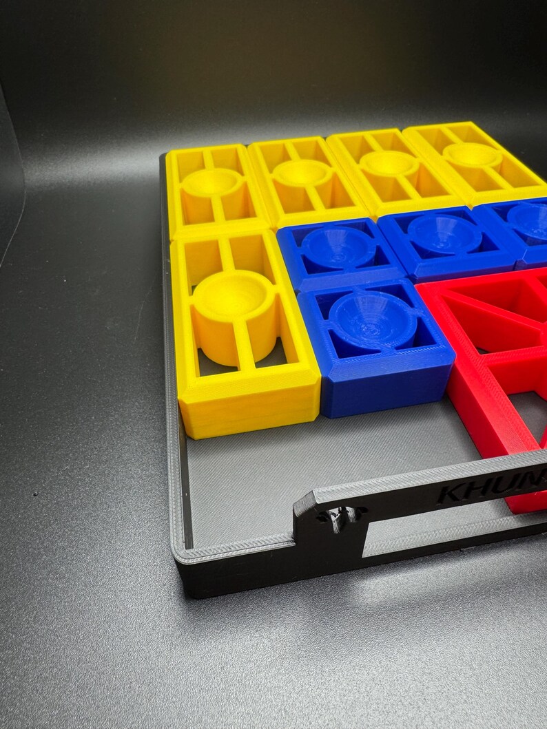 Puede incluir: Un tablero de juego negro con piezas de juego amarillas, azules y rojas. El tablero tiene un asa en el lateral y la palabra "KHUN" impresa en la parte delantera.