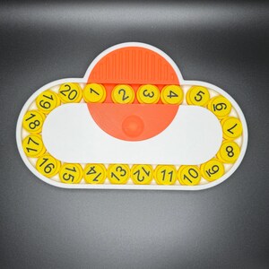 May include: A white and orange plastic toy with a circular track of yellow numbered pieces. The numbers 1 through 20 are visible on the track. The toy is designed to help children learn to count.