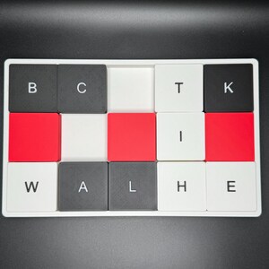 May include: A white tray with a grid of square tiles. The tiles are black, red, and white, with letters spelling "BACK THE WALL". The letters are in a bold, sans-serif font. The background is black.