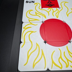 May include: A puzzle game featuring white tiles with yellow flame designs and a red sun. One tile reads "THE SUN". The game is set in a black frame.