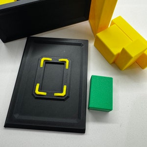 Pu&ograve; includere: Una scatola nera con una cornice gialla e un blocco verde. La scatola &egrave; aperta e l'interno &egrave; vuoto. La scatola ha una cornice gialla attorno all'apertura. Il blocco verde &egrave; appoggiato su una superficie bianca.