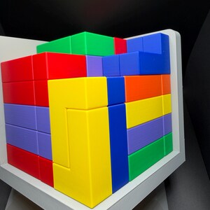 May include: A multi-colored 3D puzzle cube composed of interlocking geometric blocks in vibrant shades of red, yellow, blue, green, purple, and orange. The puzzle is housed within a white frame, displaying a complex arrangement of shapes.