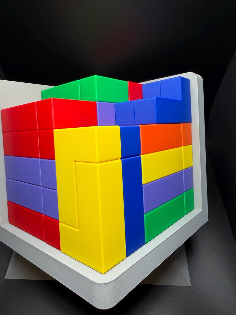 May include: A multi-colored 3D puzzle cube composed of interlocking geometric blocks in vibrant shades of red, yellow, blue, green, purple, and orange. The puzzle is housed within a white frame, displaying a complex arrangement of shapes.