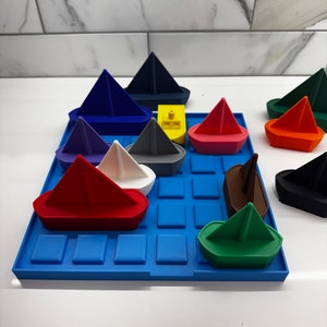 May include: A blue tray with a grid pattern holds a variety of colorful toy boats. The boats are made of plastic and are shaped like sailboats. The boats are arranged in a random pattern on the tray.