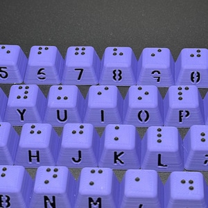 May include: Purple keyboard keys with black lettering and braille dots. The keys are arranged in a row and include the letters B, N, M, and the punctuation marks comma and period.