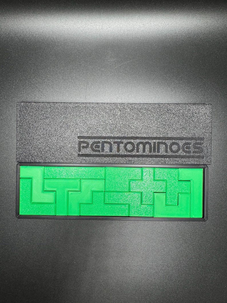 May include: A green plastic pentominoes puzzle set in a black box with the word "PENTOMINOES" printed on the lid.