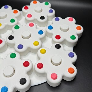 May include: White, flower-shaped toys with colorful dots around the edges. Each flower has a central, circular peg. The dots are in various colors, including red, orange, yellow, green, blue, pink, and black. The toys are on a white surface.