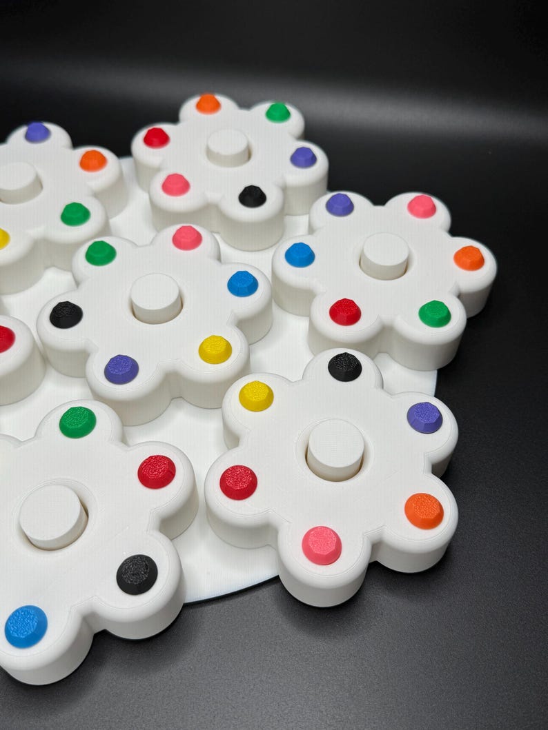 May include: White, flower-shaped toys with colorful dots around the edges. Each flower has a central, circular peg. The dots are in various colors, including red, orange, yellow, green, blue, pink, and black. The toys are on a white surface.
