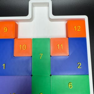 May include: A white plastic tray with a puzzle game. The puzzle pieces are numbered 1 through 13 and are colored blue, green, orange, and purple. The puzzle pieces are arranged in a grid pattern.