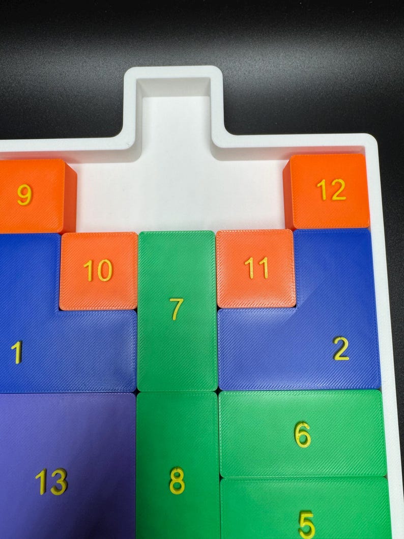 May include: A white plastic tray with a puzzle game. The puzzle pieces are numbered 1 through 13 and are colored blue, green, orange, and purple. The puzzle pieces are arranged in a grid pattern.