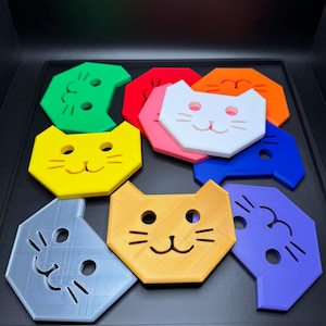 May include: A collection of colorful cat-shaped coasters in various colors, including green, red, yellow, orange, white, pink, blue, gray, and orange. Each coaster features a cat face design with eyes, nose, and whiskers. The coasters are arranged on a black surface.