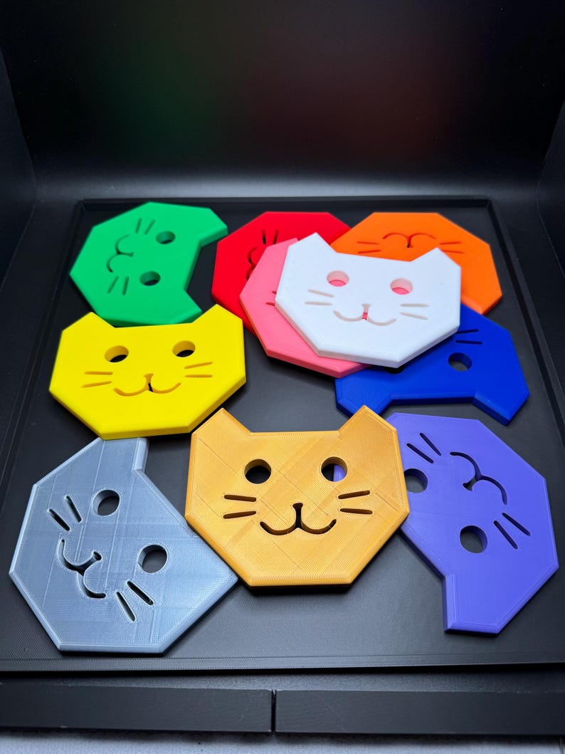 May include: A collection of colorful cat-shaped coasters in various colors, including green, red, yellow, orange, white, pink, blue, gray, and orange. Each coaster features a cat face design with eyes, nose, and whiskers. The coasters are arranged on a black surface.