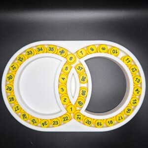 May include: A white, oval-shaped organiser tray featuring two interconnected circular compartments. The tray is lined with forty small, round, yellow tokens, each printed with a black number ranging sequentially from one to forty.