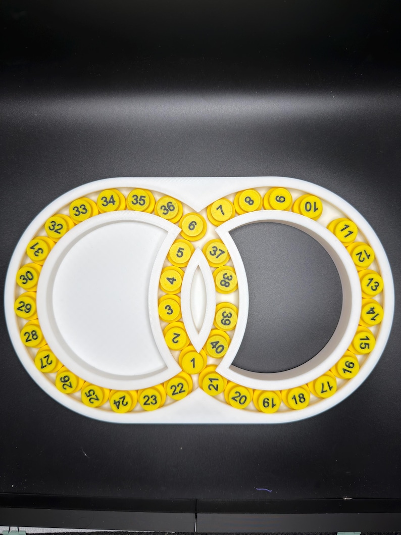 May include: A white, oval-shaped organiser tray featuring two interconnected circular compartments. The tray is lined with forty small, round, yellow tokens, each printed with a black number ranging sequentially from one to forty.