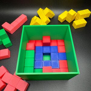 May include: A green square container filled with red, blue, and green cube-shaped blocks. Additional yellow and red blocks are scattered around the container. The blocks are arranged in a puzzle-like configuration.