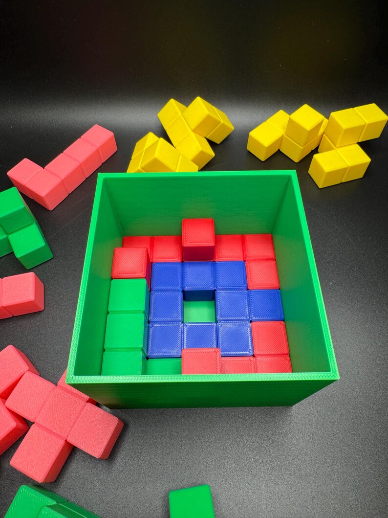 May include: A green square container filled with red, blue, and green cube-shaped blocks. Additional yellow and red blocks are scattered around the container. The blocks are arranged in a puzzle-like configuration.