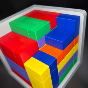 May include: A multi-colored 3D puzzle cube featuring interlocking plastic blocks in red, green, blue, yellow, orange, and purple. The puzzle is housed within a white frame, displaying a complex arrangement of geometric shapes and vibrant colors.