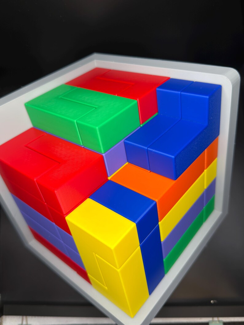 May include: A multi-colored 3D puzzle cube featuring interlocking plastic blocks in red, green, blue, yellow, orange, and purple. The puzzle is housed within a white frame, displaying a complex arrangement of geometric shapes and vibrant colors.