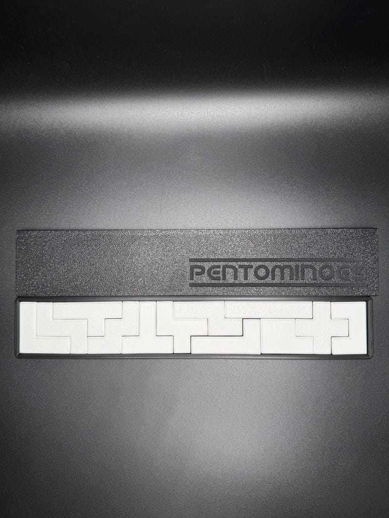May include: A black rectangular box with the word "PENTOMINO" printed on the top. The box is open and contains a white puzzle with several pieces.