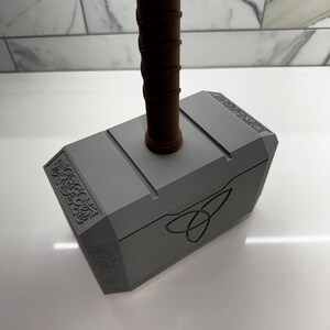 Peut inclure: Une r&eacute;plique en plastique gris du marteau de Thor, Mjolnir, avec un manche brun. Le marteau est orn&eacute; de sculptures complexes et d'un symbole de triquetra sur le devant.