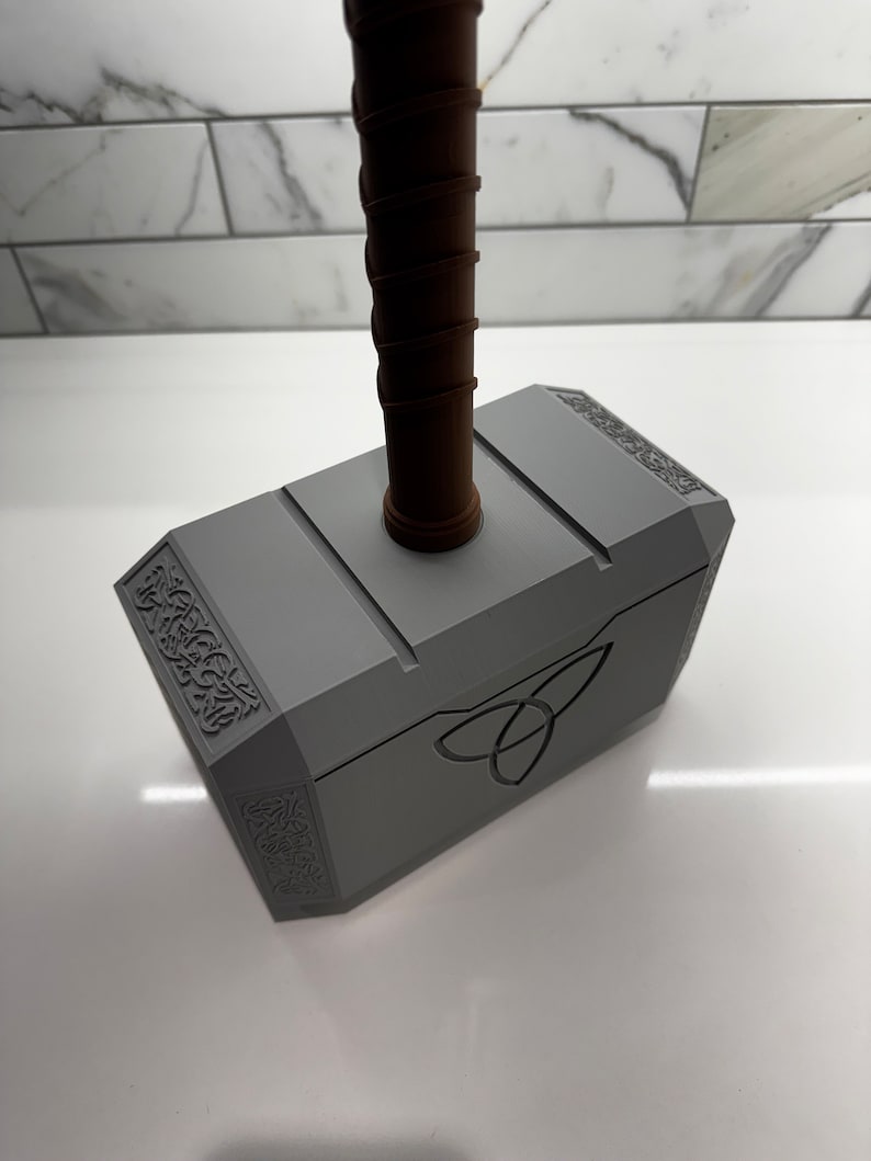 Peut inclure: Une r&eacute;plique en plastique gris du marteau de Thor, Mjolnir, avec un manche brun. Le marteau est orn&eacute; de sculptures complexes et d'un symbole de triquetra sur le devant.