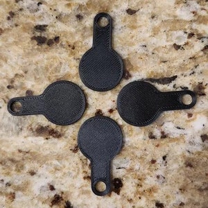 May include: Four black keyrings with a circular design and a textured finish, each with a small handle and a hole. They are arranged in a cross formation on a speckled, tan surface.