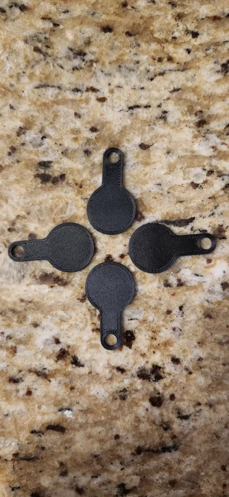 May include: Four black keyrings with a circular design and a textured finish, each with a small handle and a hole. They are arranged in a cross formation on a speckled, tan surface.
