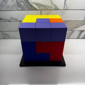 May include: A 3x3 cube puzzle made of colorful blocks. The blocks are arranged in a pattern of yellow, orange, red, blue, and purple. The cube is sitting on a black base.