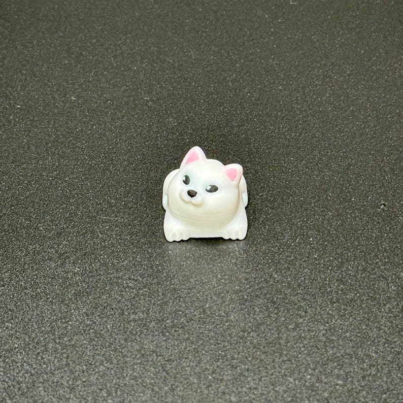 May include: A small, white, dog-shaped figurine with pink accents on the ears. The figurine has black eyes and a black nose. The dog is in a sitting position. The figurine is made of a smooth, glossy material.