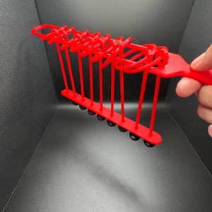 Pode incluir: Um brinquedo de pl&aacute;stico vermelho com uma s&eacute;rie de la&ccedil;os e bolas pretas que balan&ccedil;am para frente e para tr&aacute;s. O brinquedo &eacute; segurado na m&atilde;o.