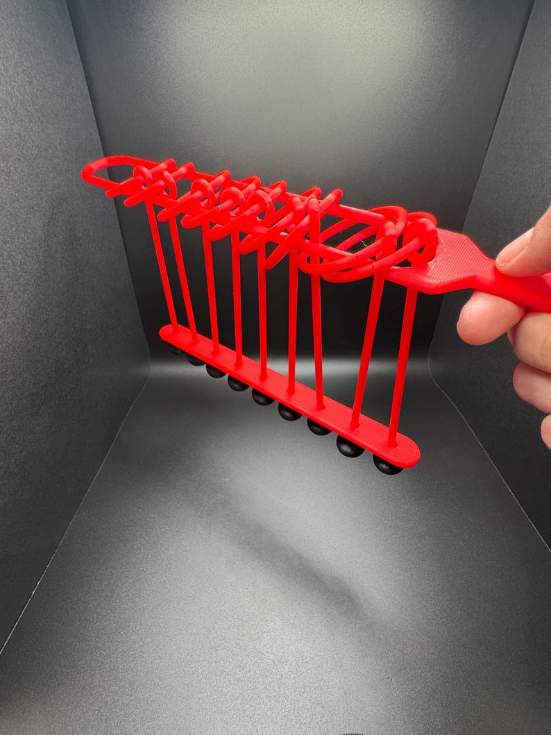 Pode incluir: Um brinquedo de pl&aacute;stico vermelho com uma s&eacute;rie de la&ccedil;os e bolas pretas que balan&ccedil;am para frente e para tr&aacute;s. O brinquedo &eacute; segurado na m&atilde;o.