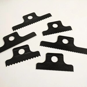 May include: Six black plastic tools with a serrated edge and a hole in the center. The tools are designed for scraping and removing materials.