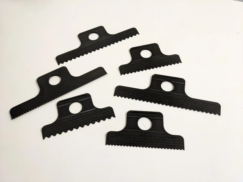 May include: Six black plastic tools with a serrated edge and a hole in the center. The tools are designed for scraping and removing materials.