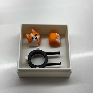 Op de afbeelding: Een witte doos bevat twee oranje en witte corgi-vormige keycaps en een zwarte keycap-trekker. E&eacute;n keycap toont het gezicht van de corgi en de andere zijn achterwerk. De keycap-trekker is zwart.